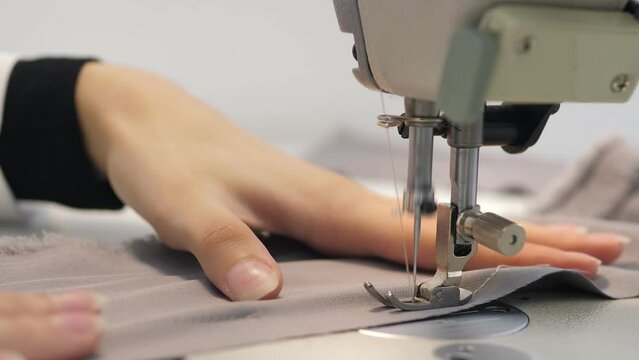 Close up slow video of woman's hand sews grey fabric on a professional sewing machine at the workplace. The seamstress's hands hold textiles for the production of dresses. Light blurred background.