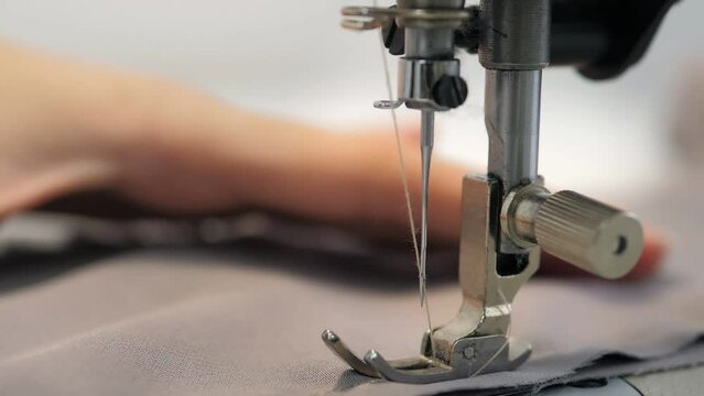 Close up slow video of woman's hand sews grey fabric on a professional sewing machine at the workplace. The seamstress's hands hold textiles for the production of dresses. Light blurred background.