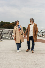 full length of cheerful senior couple in coats holding hands and walking on bridge near guard rail.