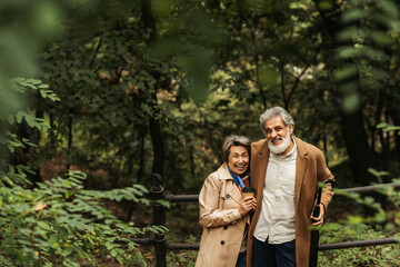 happy senior man holding umbrella and standing near wife with paper cup in park.
