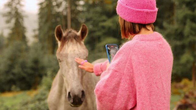 Woman in pink sweater and hat photographing on phone and cares horse that grazing on meadow in mountains. Travel and rest on nature, wildlife and rural lifestyle concept