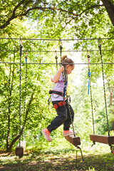 Naklejka na ściany i meble Portrait of brave little girl walk on a rope bridge in an adventure rope park. Having fun at adventure park. Scout practicing rappelling