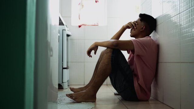 Depressed hispanic young man sitting on kitchen floor covering face in shame. Desperate South American person during difficult hard times suffering from mental illness
