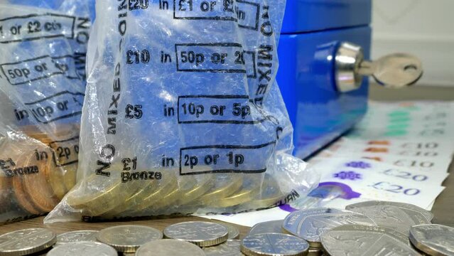 Closeup of a cash box and sterling notes, with fingers dropping two one pound coins into a plastic money bag.