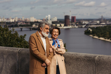 happy senior couple in beige coats smiling and walking on bridge near river.