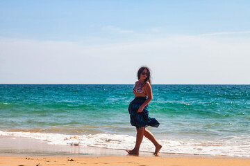 Adult woman in blue beachwear and sunglasses walking, looking away on azure sea, resting on tropical sandy beach at waves ocean background. Travel tourism vacation concept. Copy text space