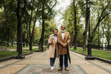 Obraz premium full length of senior woman holding paper cup while standing with stylish husband in coat.