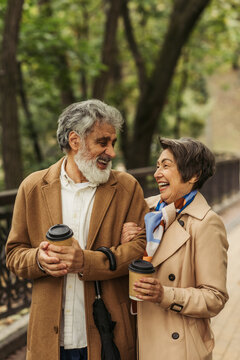 joyful retired couple in beige coats holding paper cups and walking in park.