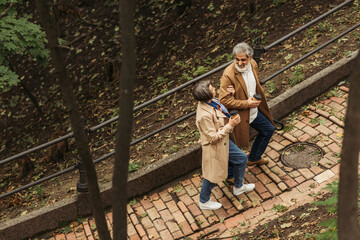 high angle view of cheerful senior couple in beige coats holding paper cups while walking in park.