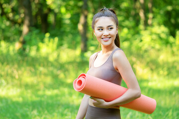 Yoga outdoors, fitness training on fresh air. Beautiful asian woman holding fitness mat, preparing for yoga practise in a forest meadow.