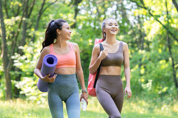 Two beautiful asian women walking with sports mats and preparing to start yoga training outdoors in a meadow.