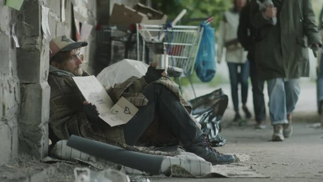Tracking shot of homeless beggar sitting on street with cardboard sign and shaking metal can while begging for money