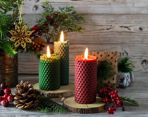 Tall candles made of natural red and green wax on wooden stands. Gift boxes and spruce branches in the background. Light wooden background