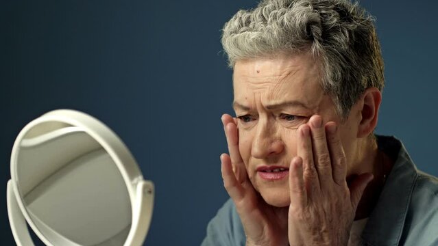 An elderly gray-haired woman carefully examines her reflection in the mirror, lightly touching the wrinkles on the skin of her face. Woman is upset.