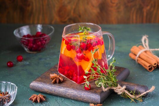 Hot autumn drink, cranberry tea with spices and orange slices in a transparent glass teapot on a wooden board on a green beon background. Hot drinks recipe. Thanksgiving Day