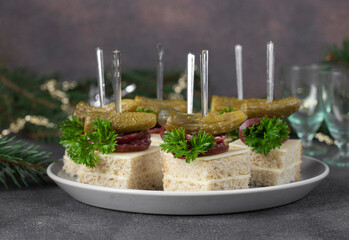 Canape with gherkins and smoked sausage on white bread with cheese on gray background, festive appetizer