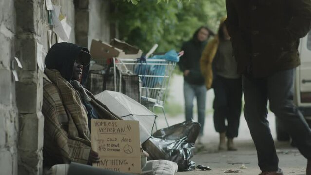 African American street beggar sitting on ground with cardboard sign and metal cup as passer by giving him money and telling kind words to support