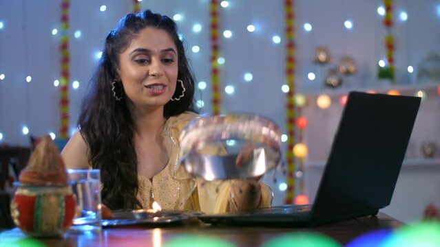 A young woman is performing online Karwa Chauth Aarti with a Puja Thali - digital world  Karwa-Chauth celebrations. A pretty lady using a sieve completes fast for her husband's long life - drinking...