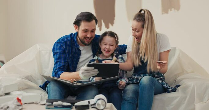 Beautiful blonde woman wearing white t-shirt and denim overalls hugs 8-year-old daughter. Girls laugh out loud as meanwhile middle-aged man tries to show them project of new apartment.