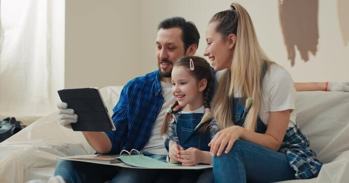 Wonderful young family is talking on online conference with friends. Brunette man holding tablet in hand and hold a folder with color palette on lap to choose the best color option for apartment.