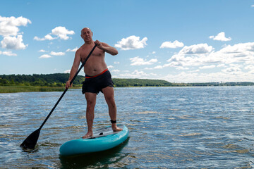 A man in shorts and a T-shirt on a SUP board swims in the lake on a sunny day against a sky with clouds.