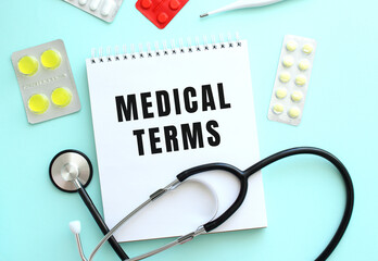 The text MEDICAL TERMS is written on a white notepad that lies next to the stethoscope and pills on a blue background.