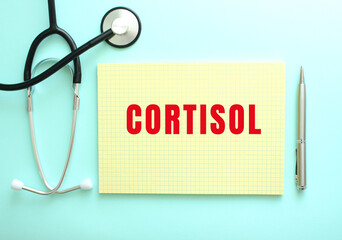 The red text CORTISOL is written in a yellow pad that lies next to the stethoscope on a blue...
