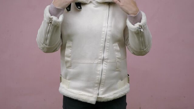 The girl zips up a light autumn jacket made of faux fur, posing directly for the camera. A woman in casual clothes against a pink wall.