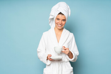 Portrait of woman in bathrobe and towel on head standing on blue background with cup of coffee