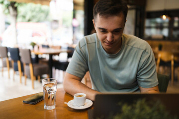 A young guy using his computer in cafe and talking on the phone while drinking water and coffee 