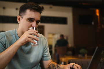 A young guy using his computer in cafe and talking on the phone while drinking water and coffee 