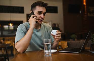 A young guy using his computer in cafe and talking on the phone while drinking water and coffee 