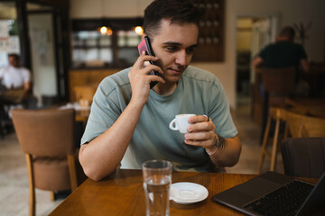 A young guy using his computer in cafe and talking on the phone while drinking water and coffee 