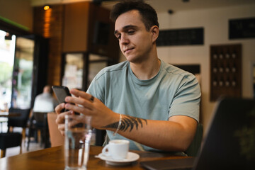 A young guy using his computer in cafe and talking on the phone while drinking water and coffee 