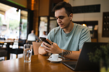 A young guy using his computer in cafe and talking on the phone while drinking water and coffee 