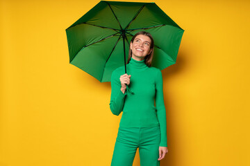Portrait of woman in green clothes holding umbrella on yellow background, looking up on clouds