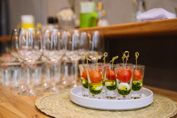 Caprese canapés in shot glasses on a skewer. cherry tomatoes, mozzarella cheese balls, pesto sauce. table with appetizers and empty wine glasses in the background.