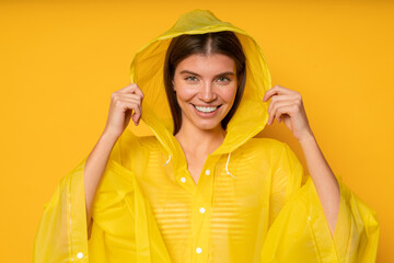Portrait of woman putting on waterproof raincoat to hide from heavy rain on yellow background
