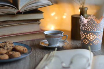 Plate of chocolate chip cookies, stack of vintage books, reading glasses, cup of tea or coffee, lit candle and fairy lights. Hygge at home. Selective focus.