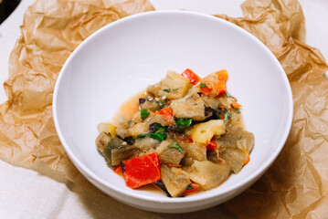 Eggplant stew with vegetables. Large cubes of stewed vegetables in a deep white ceramic bowl. View from slightly above. The dish is served hot. Ingredients: cauliflower, red bell peppers, parsley.