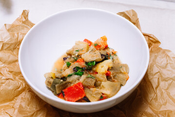 Eggplant stew with vegetables. Large cubes of stewed vegetables in a deep white ceramic bowl. View from slightly above. The dish is served hot. Ingredients: cauliflower, red bell peppers, parsley.