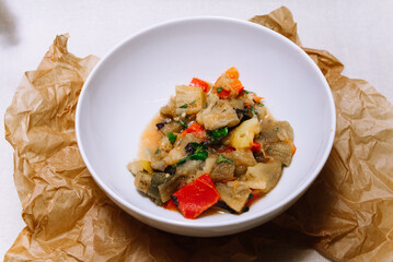 Eggplant stew with vegetables. Large cubes of stewed vegetables in a deep white ceramic bowl. View from slightly above. The dish is served hot. Ingredients: cauliflower, red bell peppers, parsley.