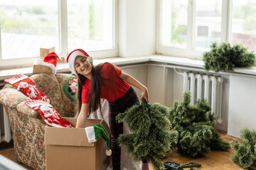little girl makes an artificial Christmas tree wear santa hat indoors - obrazy, fototapety, plakaty
