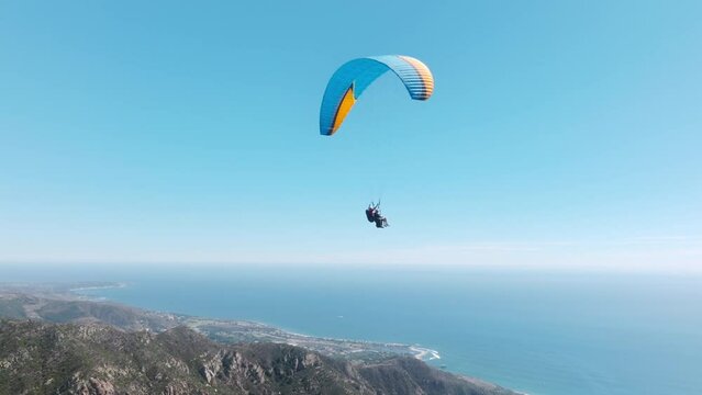 Slow motion up in the air, cinematic aerial drone footage above Malibu USA. Extreme paraglider flying against a clear blue sky, sunbeam shines into camera. Man with instructor flies with paraglide 4k