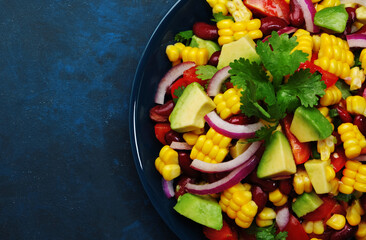 Mexican spicy salad with corn, red beans, avocado, jalapeno peppers, cherry tomatoes, onion and cilantro. Blue table background, top view