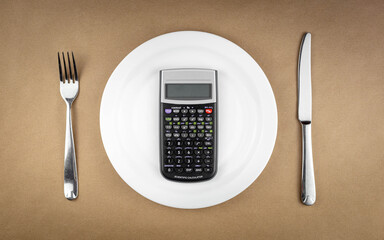 Diet concept with calculator on empty plate with fork and knife isolated on brown recycled paper background with copy space, food spending costs concept