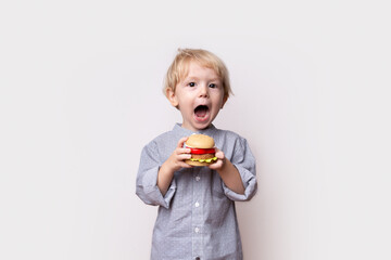 little toddler boy with blond hair in a shirt holds a toy plastic hamburger in his hands on a white...