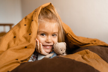 cute little girl lies cozy in bed covered with a blanket over her head, smiling, with copy space