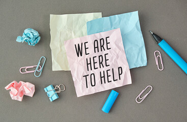 WE ARE HERE TO HELP text in notebook on gray background next to pencils, pen and paper clips