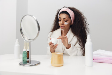 Aesthetic medicine skincare beauty products ad concept. Flawless cheerful tanned curly Latin lady put facial cream on cheeks enjoying admiring herself smiling in mirror at home interior. Copy space